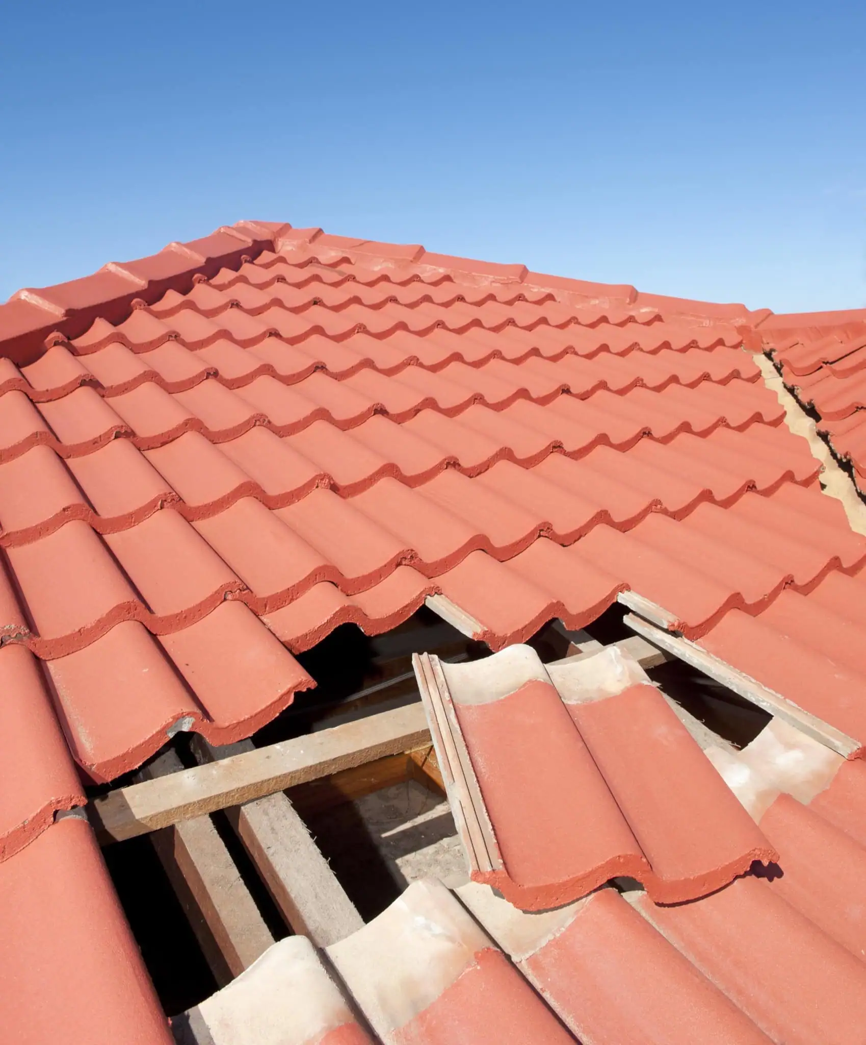 A detailed, close-up shot of a red-tiled roof with a section of missing and damaged tiles, revealing the wooden support structure underneath. The tiles are a wavy design, and the sky above is a clear blue.