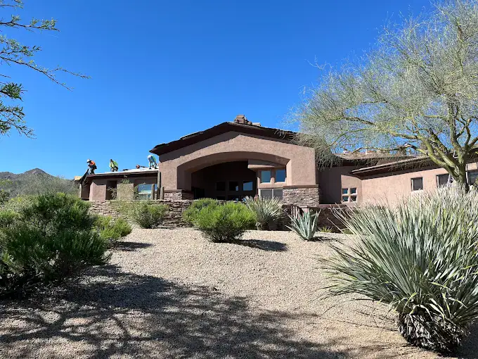 A southwestern-style house with a tan exterior, surrounded by desert landscaping and shrubs, with workers visible on the roof under a clear blue sky.