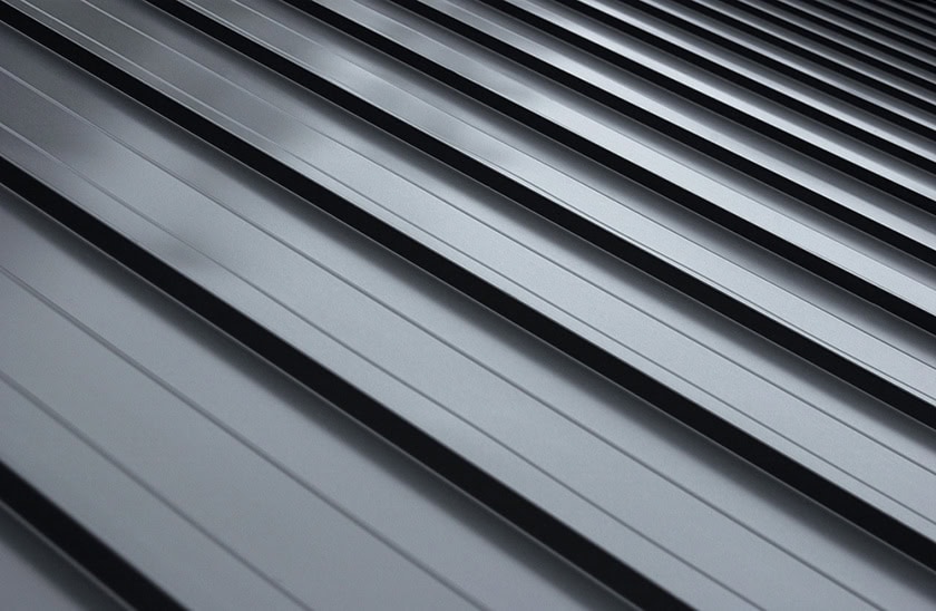 Close-up view of a sleek, gray metal roof with parallel raised ridges, creating a pattern of alternating shadows and highlights. The surface appears clean and modern, with a diagonal perspective.