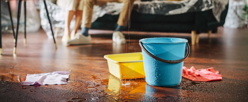 A blue bucket and a yellow tub catch water from a leak on a wooden floor, surrounded by wet cloths. In the background, two people sit on a sofa, mostly out of focus.
