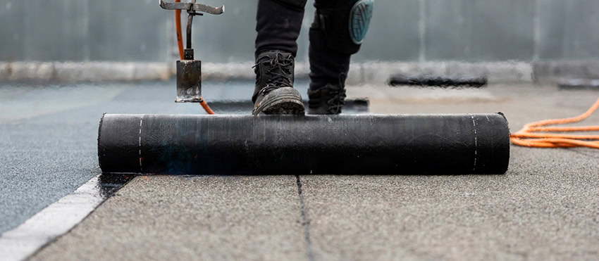 Close-up of a worker's legs as they unroll and install a black roofing membrane on a flat roof, using a torch to seal the material.