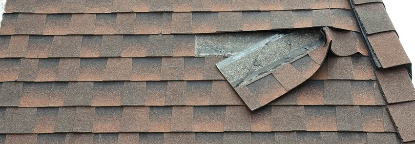 A section of brown and black asphalt roof shingles is damaged, with several shingles lifted and exposing the underlying roofing material beneath.