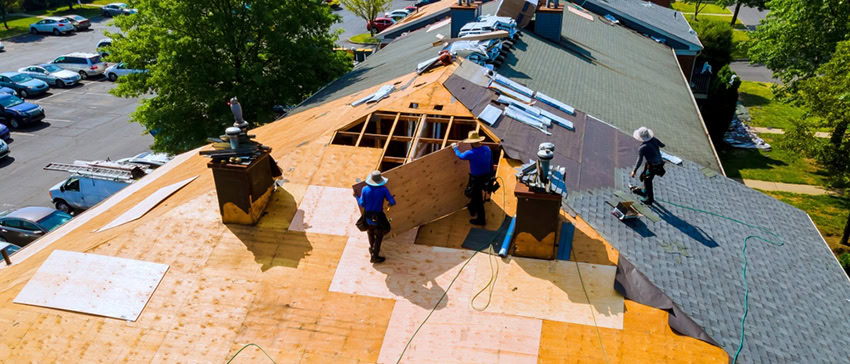 Aerial view of workers repairing and replacing shingles on a large roof, with tools and materials scattered around. Trees and a parking lot are visible in the background.