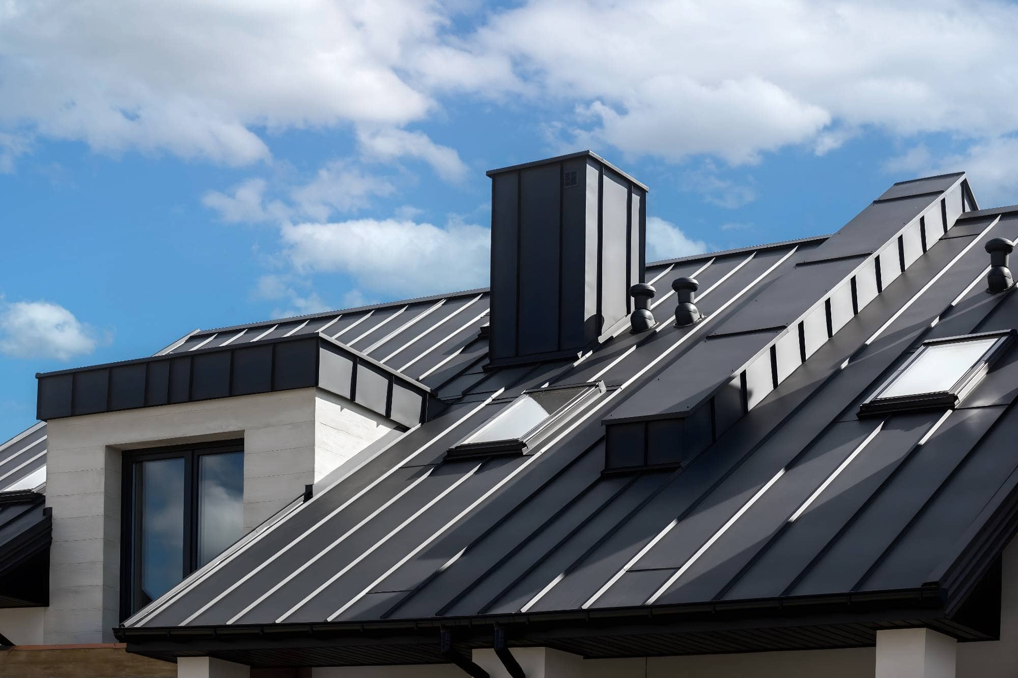Modern house with a sleek, dark metal roof featuring multiple skylights, a large chimney, and vertical standing seams—expertly installed by a top roofer Maricopa County—set against a blue sky with scattered clouds.