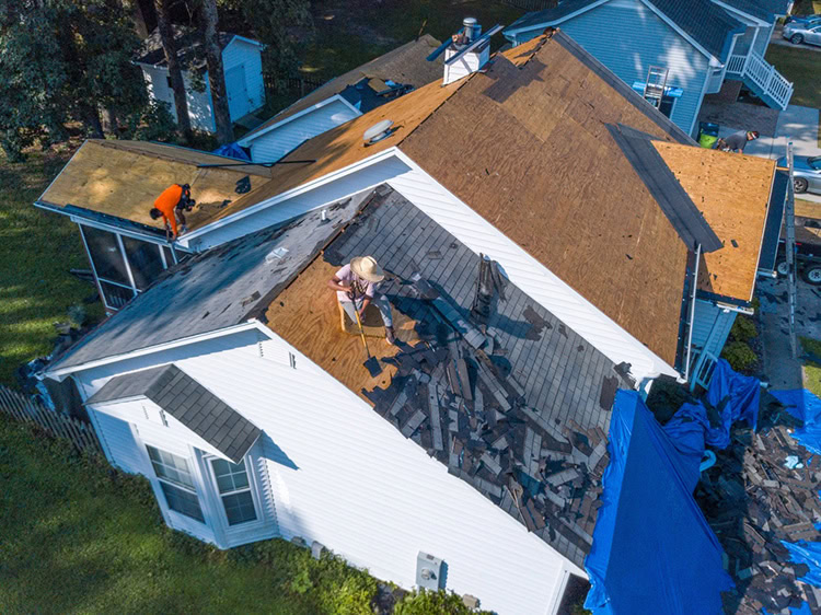 Aerial view of two workers from a roofing contractor Maricopa County, AZ, removing old shingles and repairing the roof of a white house, with debris scattered and blue tarps covering parts of the ground and house.