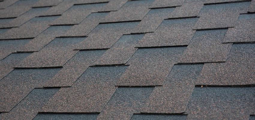 Close-up view of overlapping asphalt roof shingles arranged in a staggered pattern, showing rough texture and muted brown and gray tones.