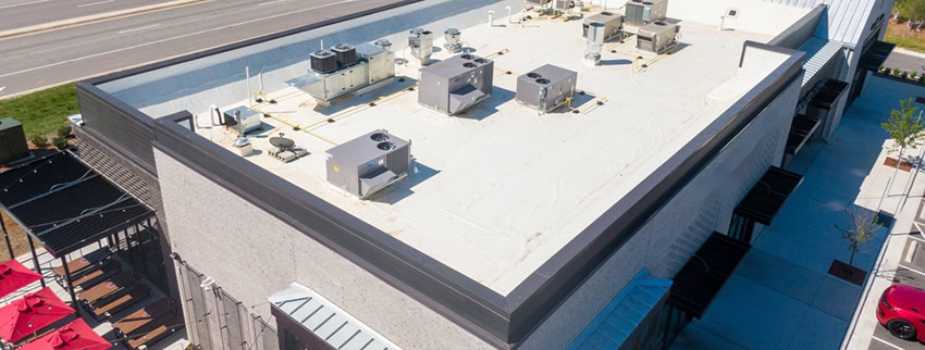 Aerial view of a commercial building with a flat white roof featuring multiple HVAC units and vents. The building is next to a road, with sidewalks, awnings, and outdoor seating areas visible below.